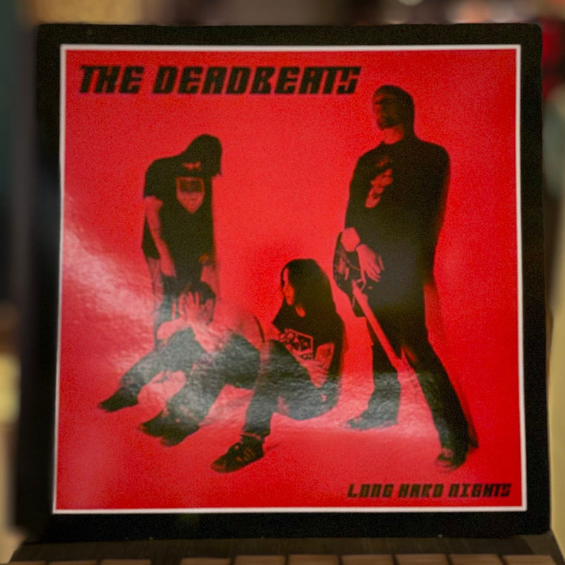 Album cover for 'The Deadbeats' with black figures on a red background, displayed on a wooden surface.