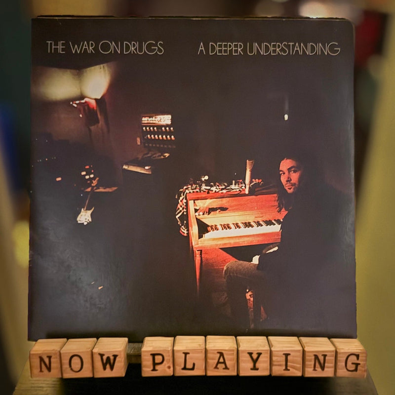 Album cover for 'The War on Drugs' with a person playing piano, text 'A Deeper Understanding', and 'Now Playing' sign.